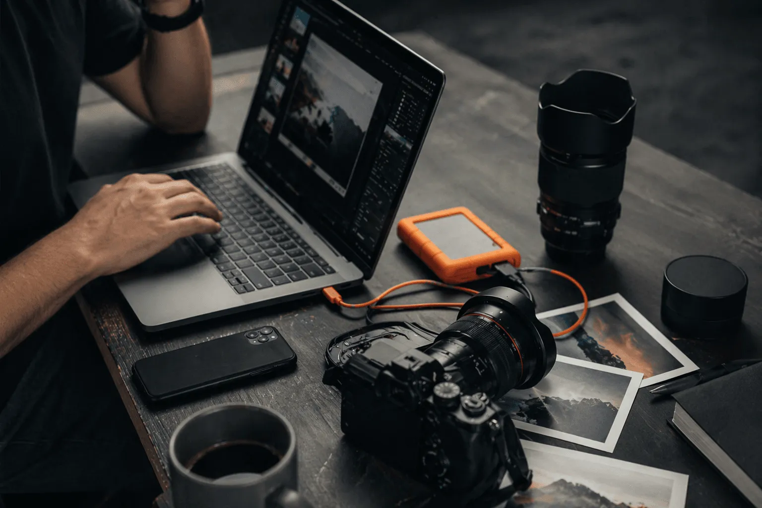 Persona editando fotos en un portátil rodeada de equipo fotográfico, como una cámara, objetivos y un disco duro